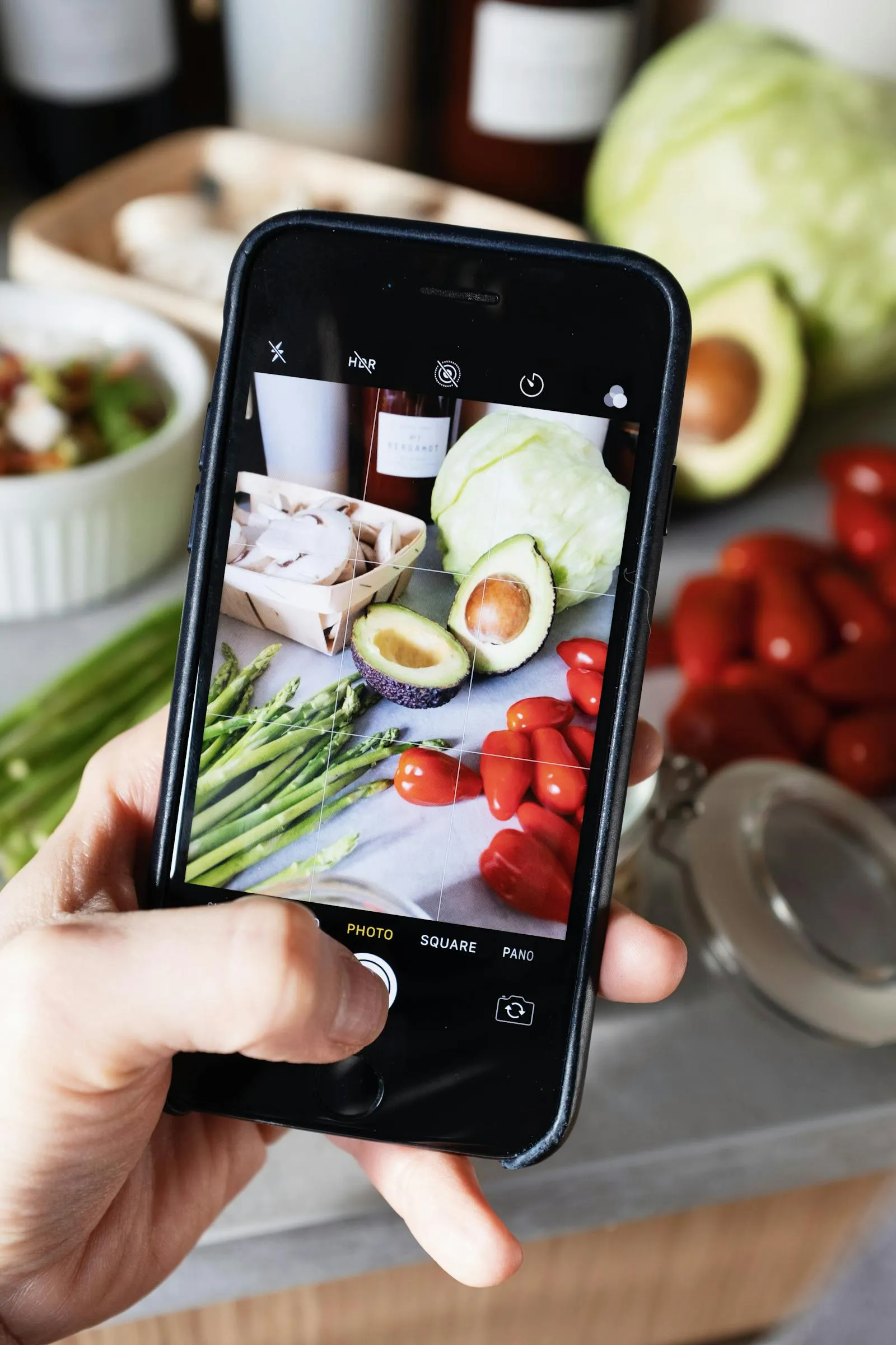 Hands holding a smartphone over fresh vegetables on a wooden table — documenting meals for AI analysis