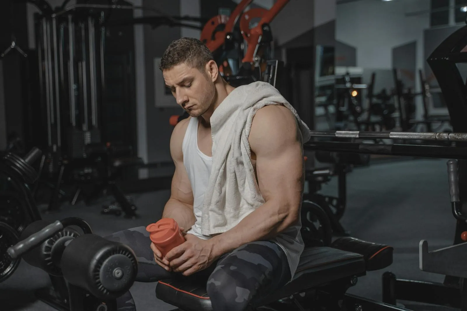A muscular athlete sits in a gym after training, holding a protein shaker bottle with a towel on his shoulder