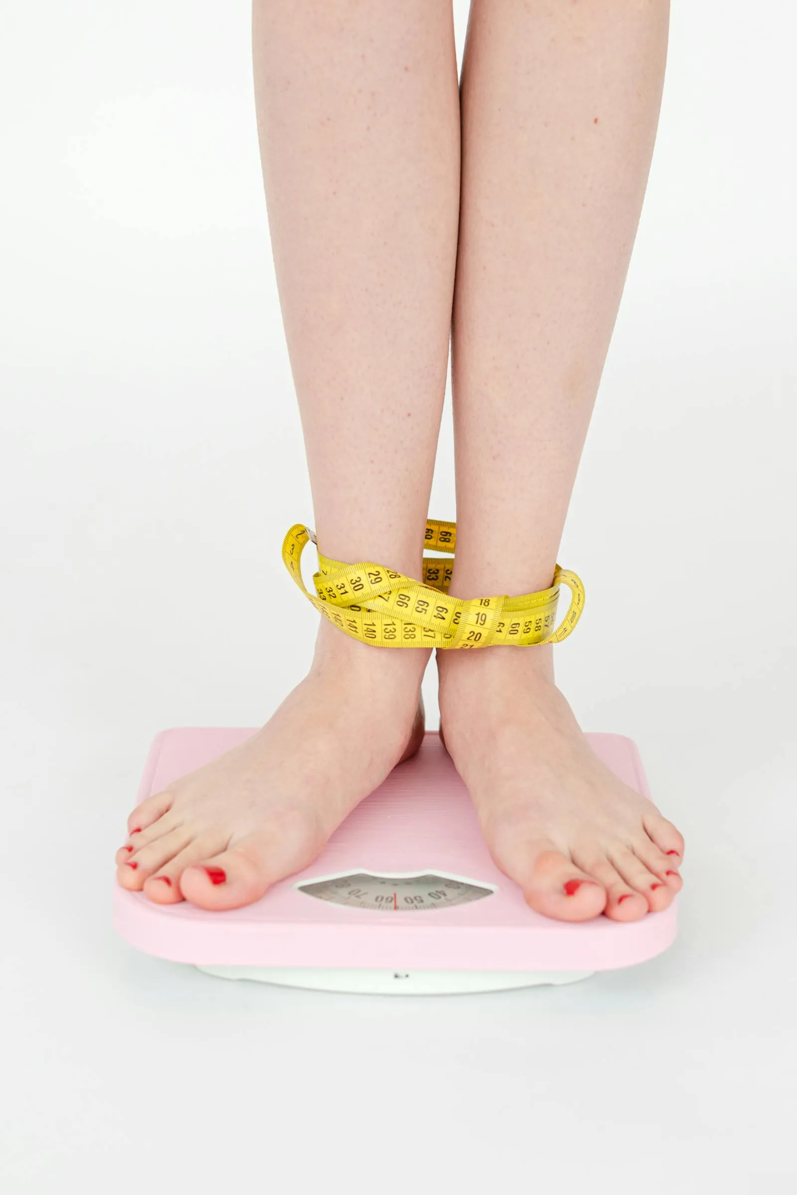 A woman holds a measuring tape around her thigh while standing on a bathroom scale — combining weight and circumference data